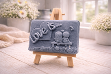 Load image into Gallery viewer, A rectangular handmade vegan soap featuring a detailed 3D relief carving of a young couple sitting on a bench under a crescent moon, with the words "Love is..." embossed inside a heart, displayed on a wooden easel.
