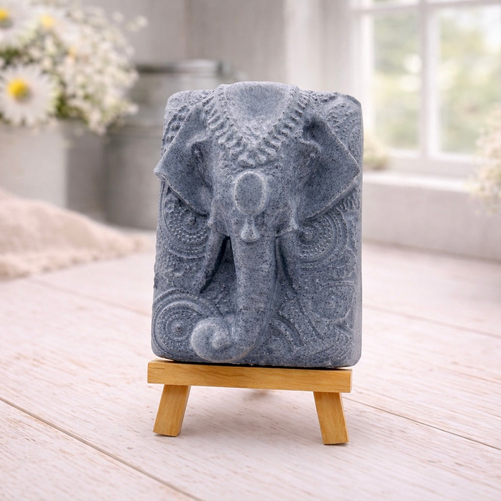 A rectangular handmade vegan soap in a speckled slate-grey finish, featuring a highly detailed relief of an elephant's head adorned with ornate, traditional patterns and a decorative floral background, displayed on a miniature wooden easel.