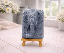 Load image into Gallery viewer, A rectangular handmade vegan soap in a speckled slate-grey finish, featuring a highly detailed relief of an elephant's head adorned with ornate, traditional patterns and a decorative floral background, displayed on a miniature wooden easel.
