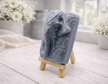Load image into Gallery viewer, A rectangular handmade vegan soap in a speckled slate-grey finish, featuring a highly detailed relief of an elephant's head adorned with ornate, traditional patterns and a decorative floral background, displayed on a miniature wooden easel.
