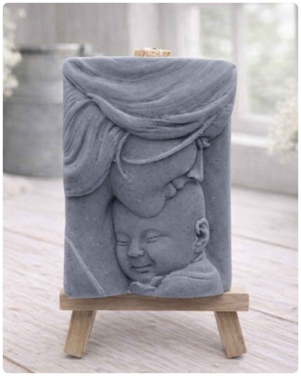 A rectangular handmade vegan soap featuring a high-relief carving of a mother tenderly cradling a small baby in her arms, displayed on a miniature wooden easel in a bright farmhouse setting.