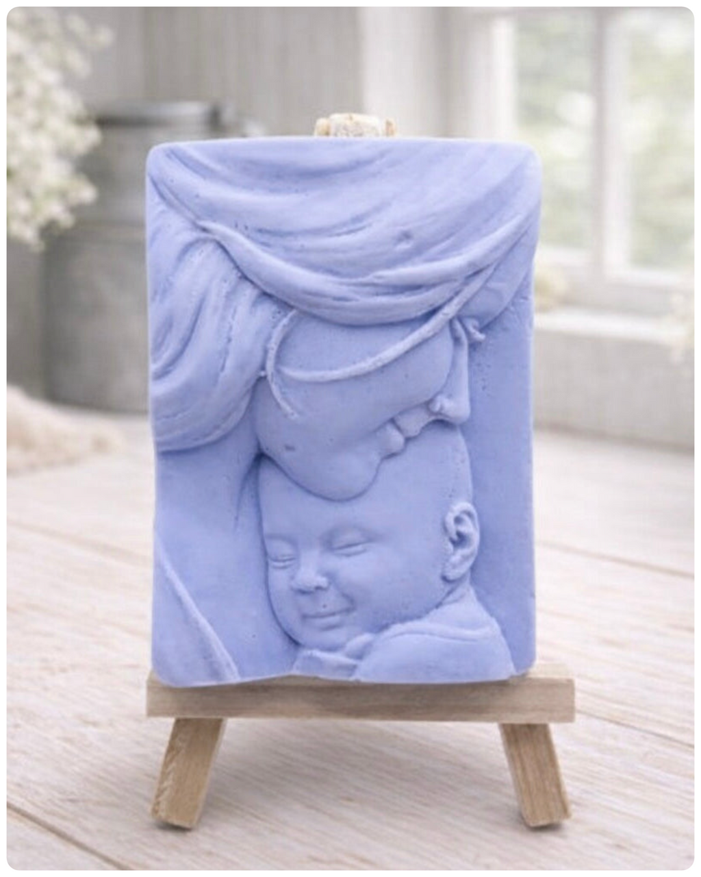 A rectangular handmade vegan soap featuring a high-relief carving of a mother tenderly cradling a small baby in her arms, displayed on a miniature wooden easel in a bright farmhouse setting.