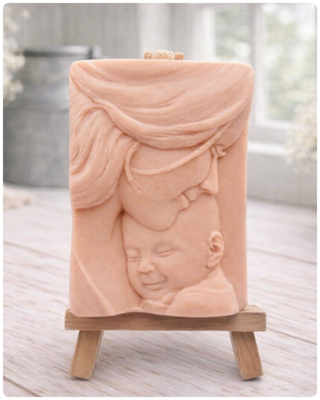A rectangular handmade vegan soap featuring a high-relief carving of a mother tenderly cradling a small baby in her arms, displayed on a miniature wooden easel in a bright farmhouse setting.