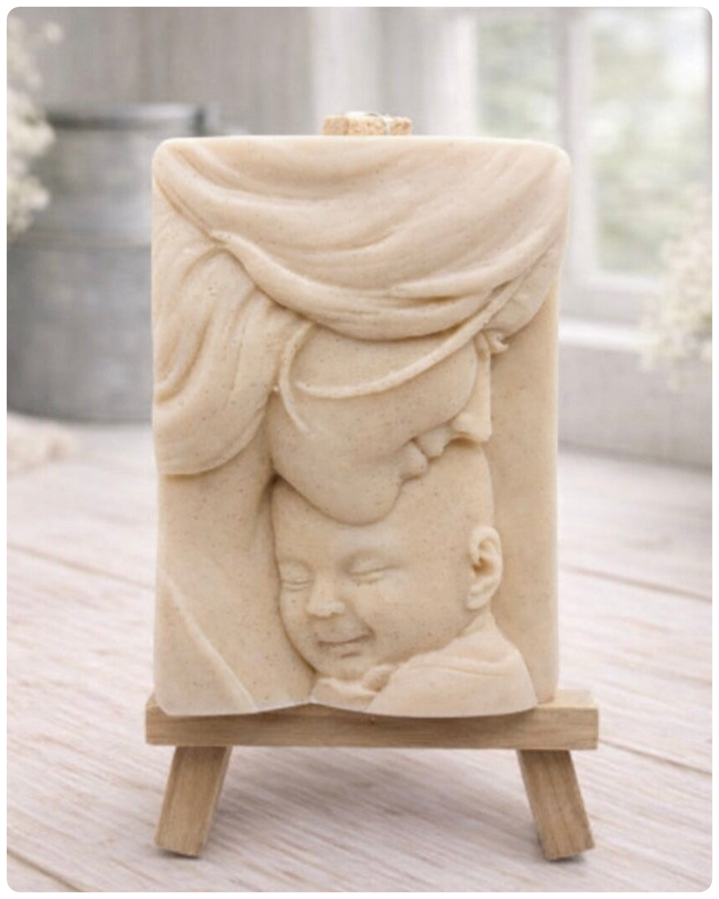 A rectangular handmade vegan soap featuring a high-relief carving of a mother tenderly cradling a small baby in her arms, displayed on a miniature wooden easel in a bright farmhouse setting.
