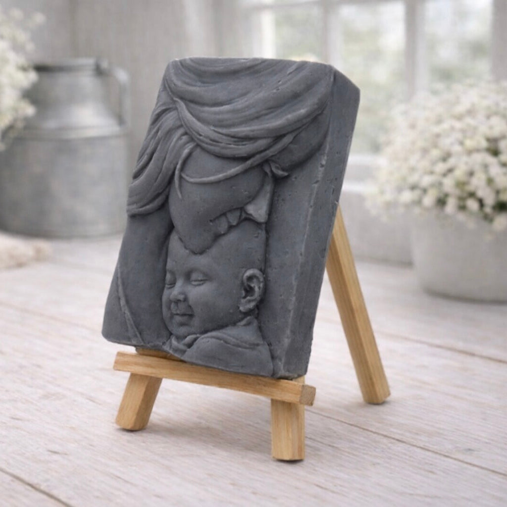 A rectangular handmade vegan soap featuring a high-relief carving of a mother tenderly cradling a small baby in her arms, displayed on a miniature wooden easel in a bright farmhouse setting.