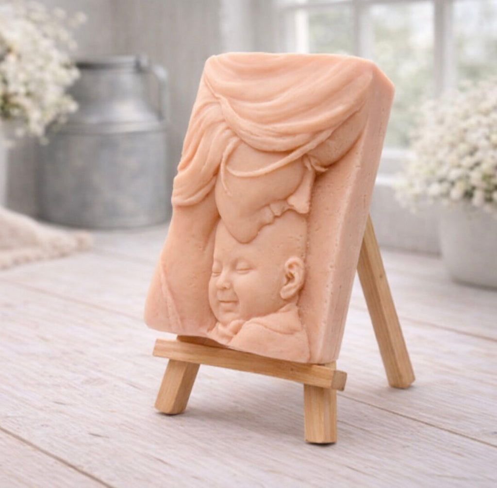 A rectangular handmade vegan soap featuring a high-relief carving of a mother tenderly cradling a small baby in her arms, displayed on a miniature wooden easel in a bright farmhouse setting.