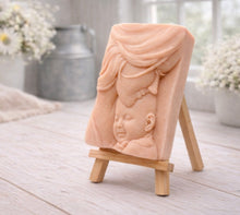 Load image into Gallery viewer, A rectangular handmade vegan soap featuring a high-relief carving of a mother tenderly cradling a small baby in her arms, displayed on a miniature wooden easel in a bright farmhouse setting.
