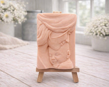Load image into Gallery viewer, A rectangular handmade vegan soap featuring a high-relief carving of a mother tenderly cradling a small baby in her arms, displayed on a miniature wooden easel in a bright farmhouse setting.
