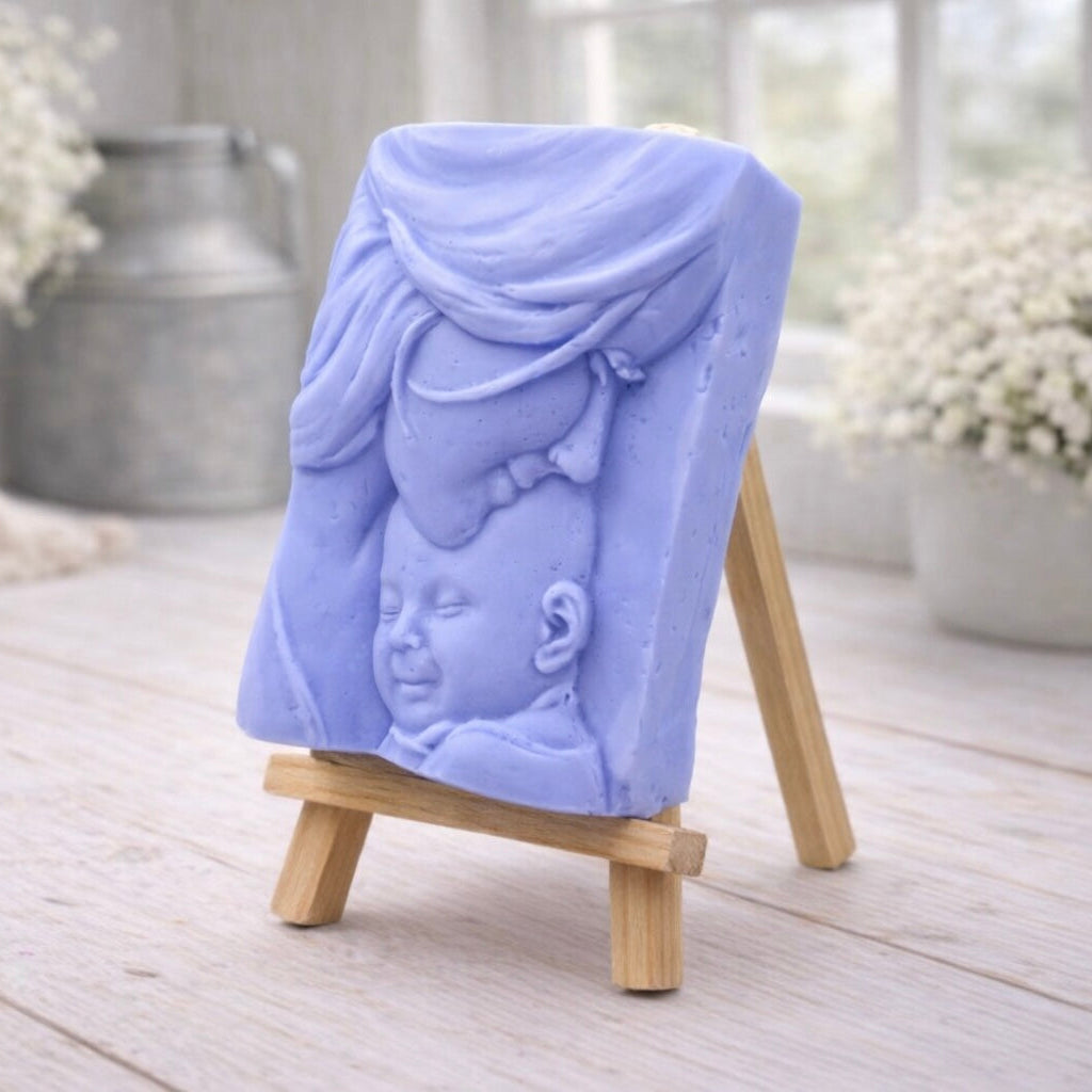 A rectangular handmade vegan soap featuring a high-relief carving of a mother tenderly cradling a small baby in her arms, displayed on a miniature wooden easel in a bright farmhouse setting.