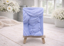 Load image into Gallery viewer, A rectangular handmade vegan soap featuring a high-relief carving of a mother tenderly cradling a small baby in her arms, displayed on a miniature wooden easel in a bright farmhouse setting.
