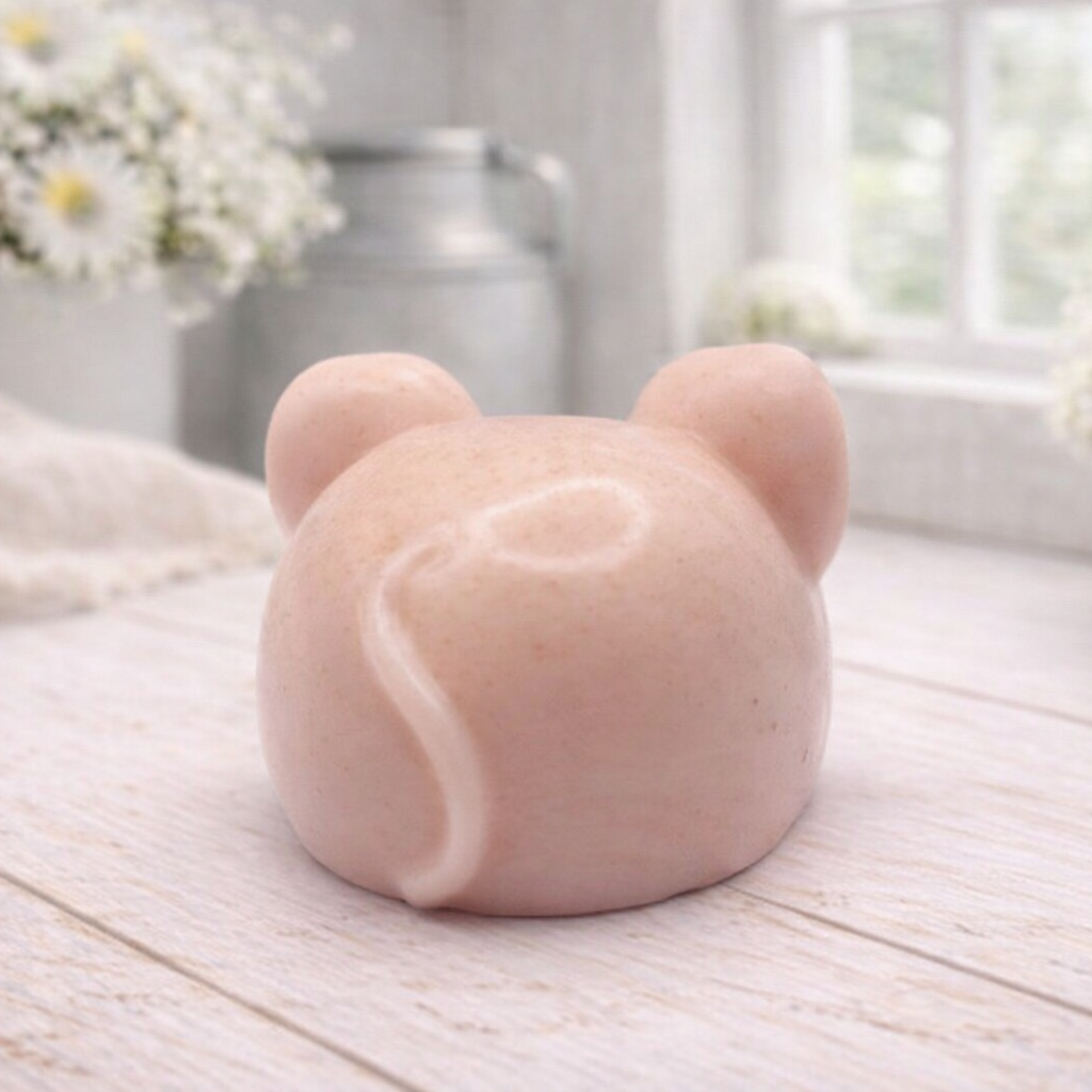 A 3D handmade vegan soap sculpted into the shape of a cute resting mouse with large rounded ears and a delicate snout, displayed on a light wooden surface in a bright farmhouse setting