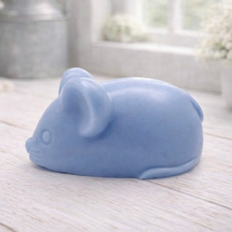 A 3D handmade vegan soap sculpted into the shape of a cute resting mouse with large rounded ears and a delicate snout, displayed on a light wooden surface in a bright farmhouse setting