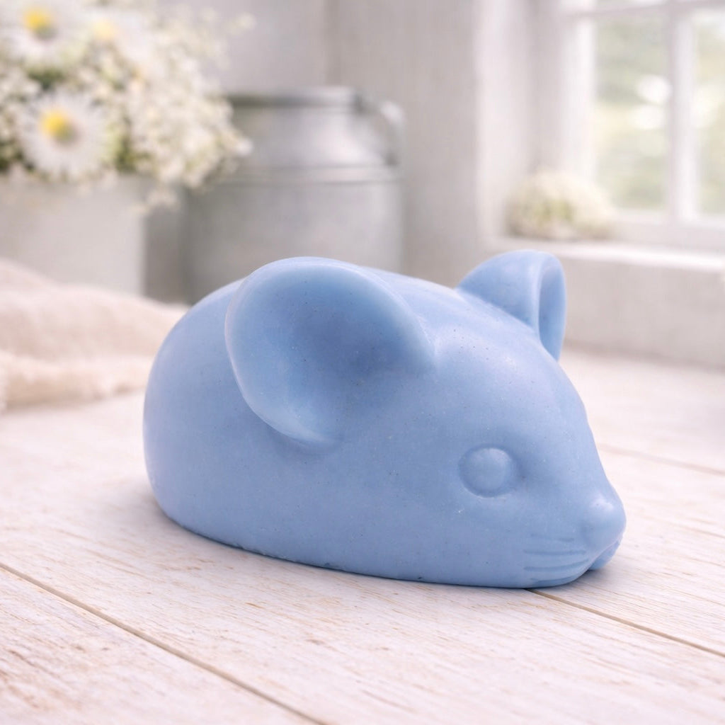 A 3D handmade vegan soap sculpted into the shape of a cute resting mouse with large rounded ears and a delicate snout, displayed on a light wooden surface in a bright farmhouse setting