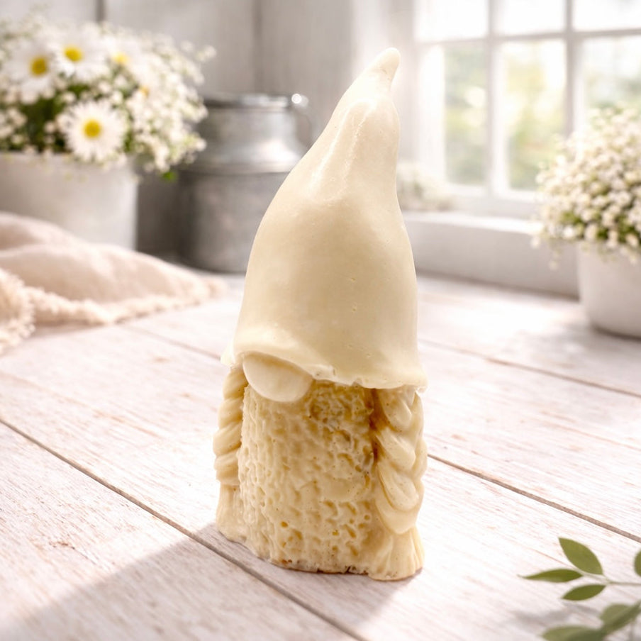 A handmade vegan soap sculpted in the shape of a female gnome (or gonk), featuring a tall, slightly curved hat pulled over the eyes and two thick, textured plaits framing a textured dress, in a natural cream colour.