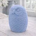 A detailed large handmade vegan soap gift in the shape of an owl with intricate feather textures, displayed on a light wooden table with white flowers in the background.