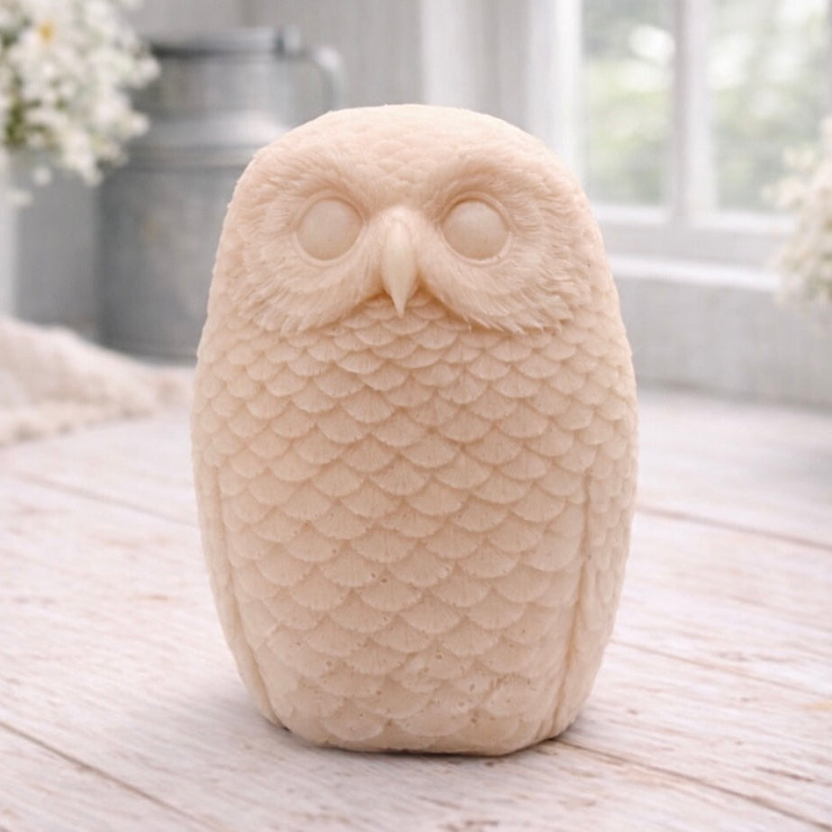 A detailed handmade vegan soap in the shape of an owl with intricate feather textures, displayed on a light wooden table with white flowers in the background.