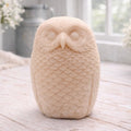 A detailed handmade vegan soap in the shape of an owl with intricate feather textures, displayed on a light wooden table with white flowers in the background.
