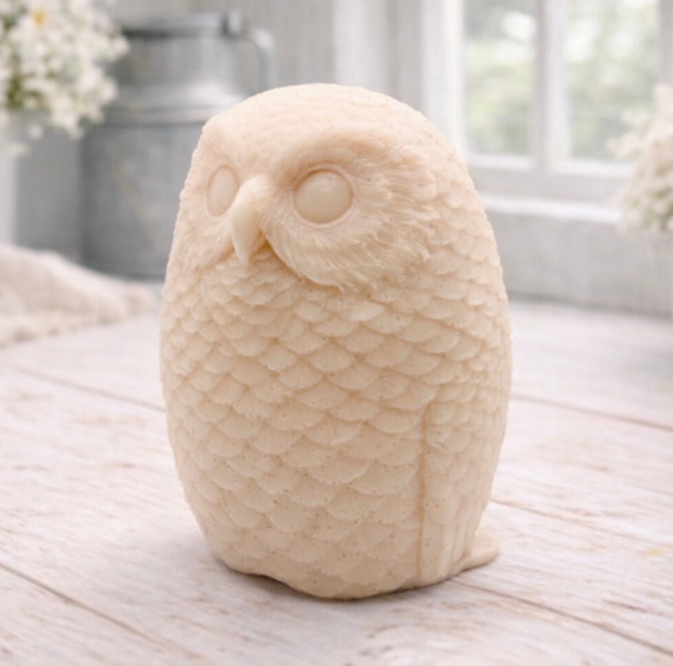 A detailed handmade vegan soap in the shape of an owl with intricate feather textures, displayed on a light wooden table with white flowers in the background.