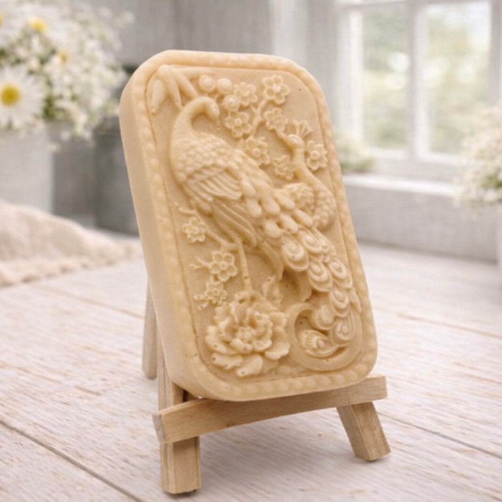 handmade vegan soap featuring a highly detailed relief carving of a peacock with a long, flowing tail, surrounded by delicate cherry blossoms and a large peony, displayed on a wooden easel.