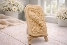 Load image into Gallery viewer, handmade vegan soap featuring a highly detailed relief carving of a peacock with a long, flowing tail, surrounded by delicate cherry blossoms and a large peony, displayed on a wooden easel.
