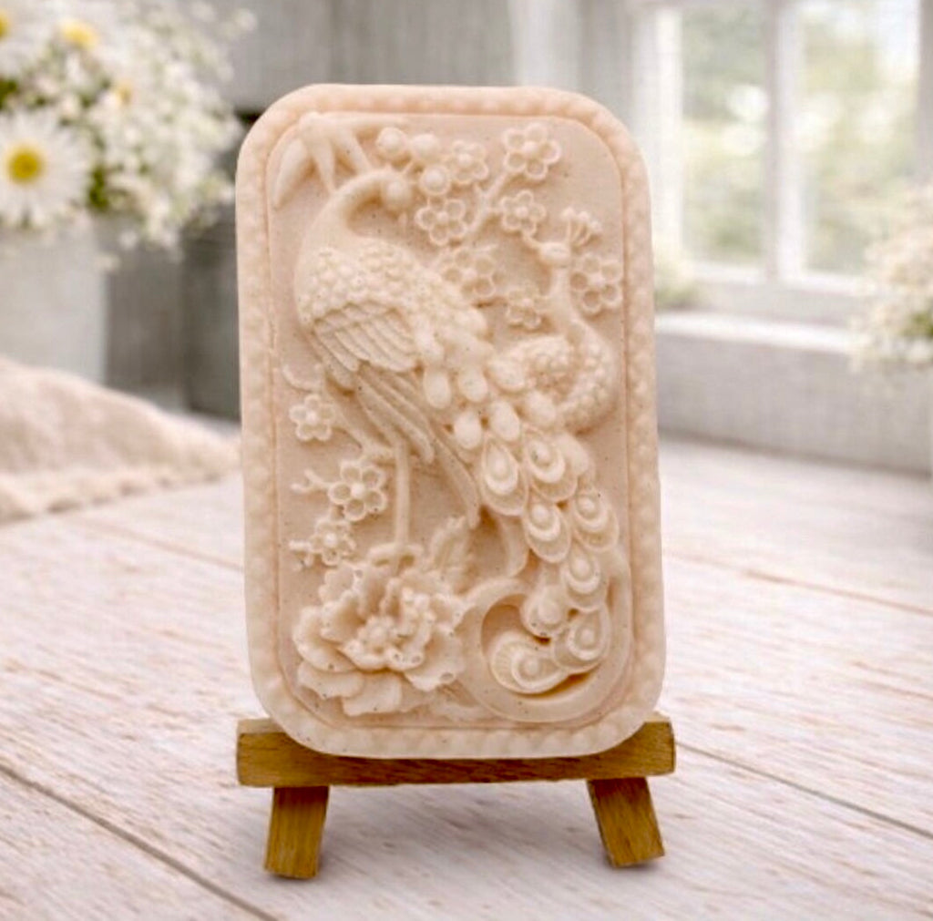 handmade vegan soap featuring a highly detailed relief carving of a peacock with a long, flowing tail, surrounded by delicate cherry blossoms and a large peony, displayed on a wooden easel.