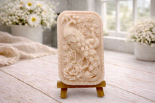 Load image into Gallery viewer, handmade vegan soap featuring a highly detailed relief carving of a peacock with a long, flowing tail, surrounded by delicate cherry blossoms and a large peony, displayed on a wooden easel.
