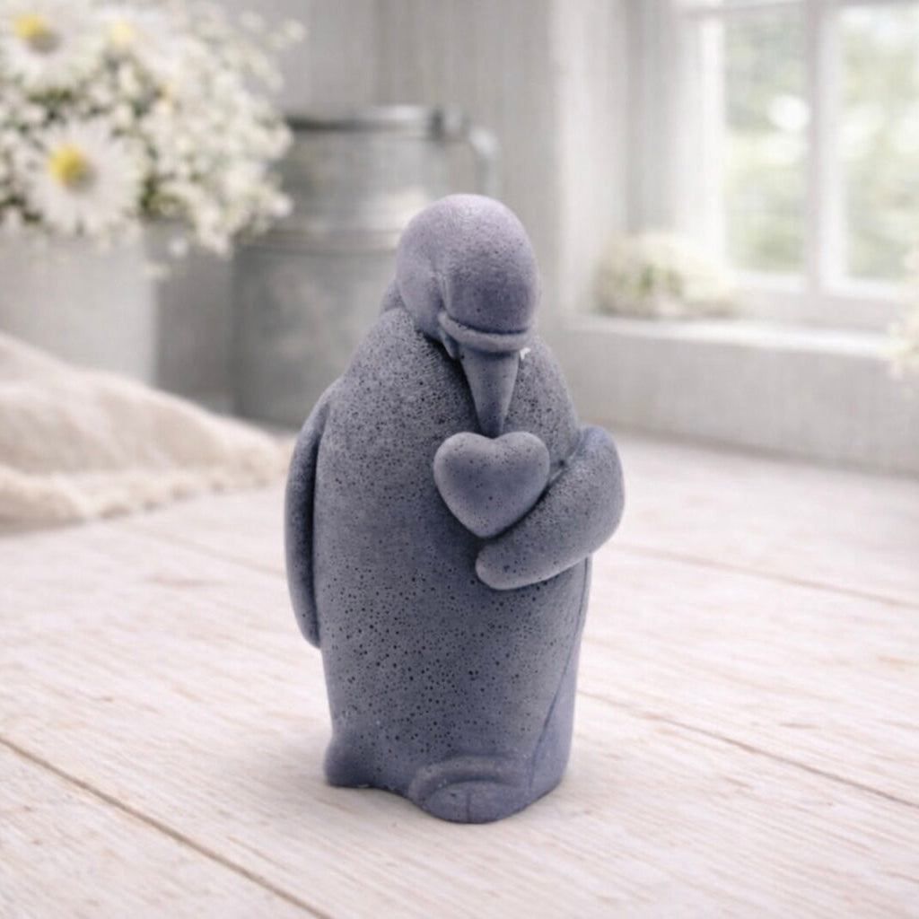 A 3D handmade vegan soap sculpted as a standing penguin tenderly cradling a heart against its chest, displayed on a light wooden surface in a bright farmhouse setting.