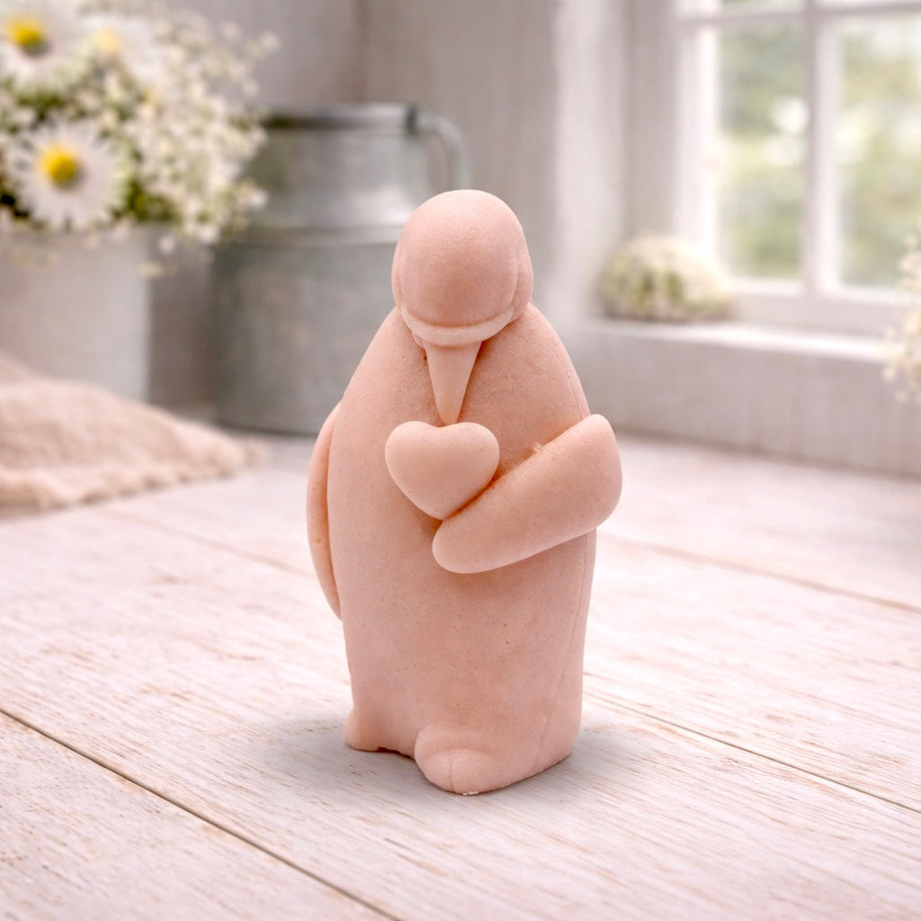 A 3D handmade vegan soap sculpted as a standing penguin tenderly cradling a heart against its chest, displayed on a light wooden surface in a bright farmhouse setting.