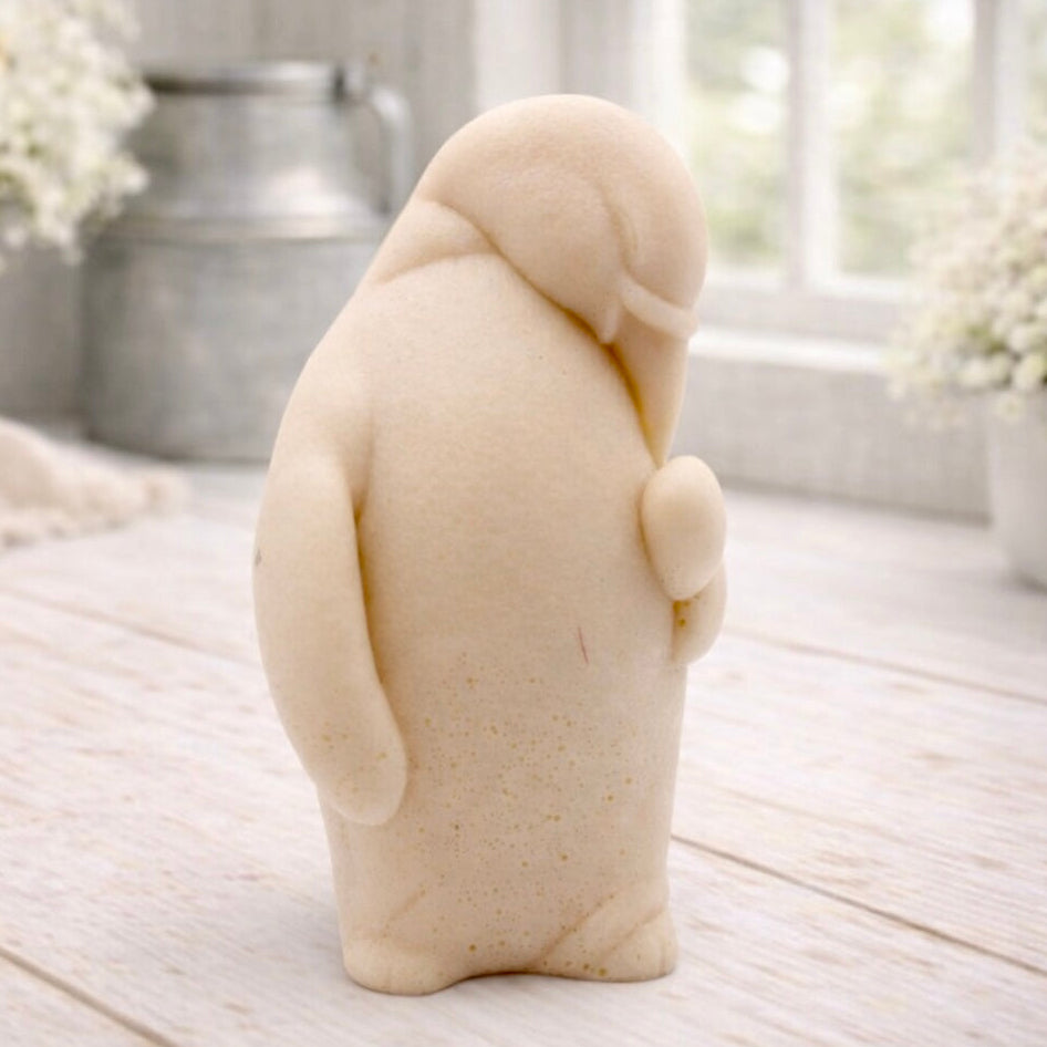 A 3D handmade vegan soap sculpted as a standing penguin tenderly cradling a heart against its chest, displayed on a light wooden surface in a bright farmhouse setting.