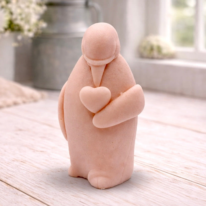 A handmade vegan soap sculpted as a standing penguin tenderly cradling a heart against its chest, displayed on a light wooden surface in a bright farmhouse setting.