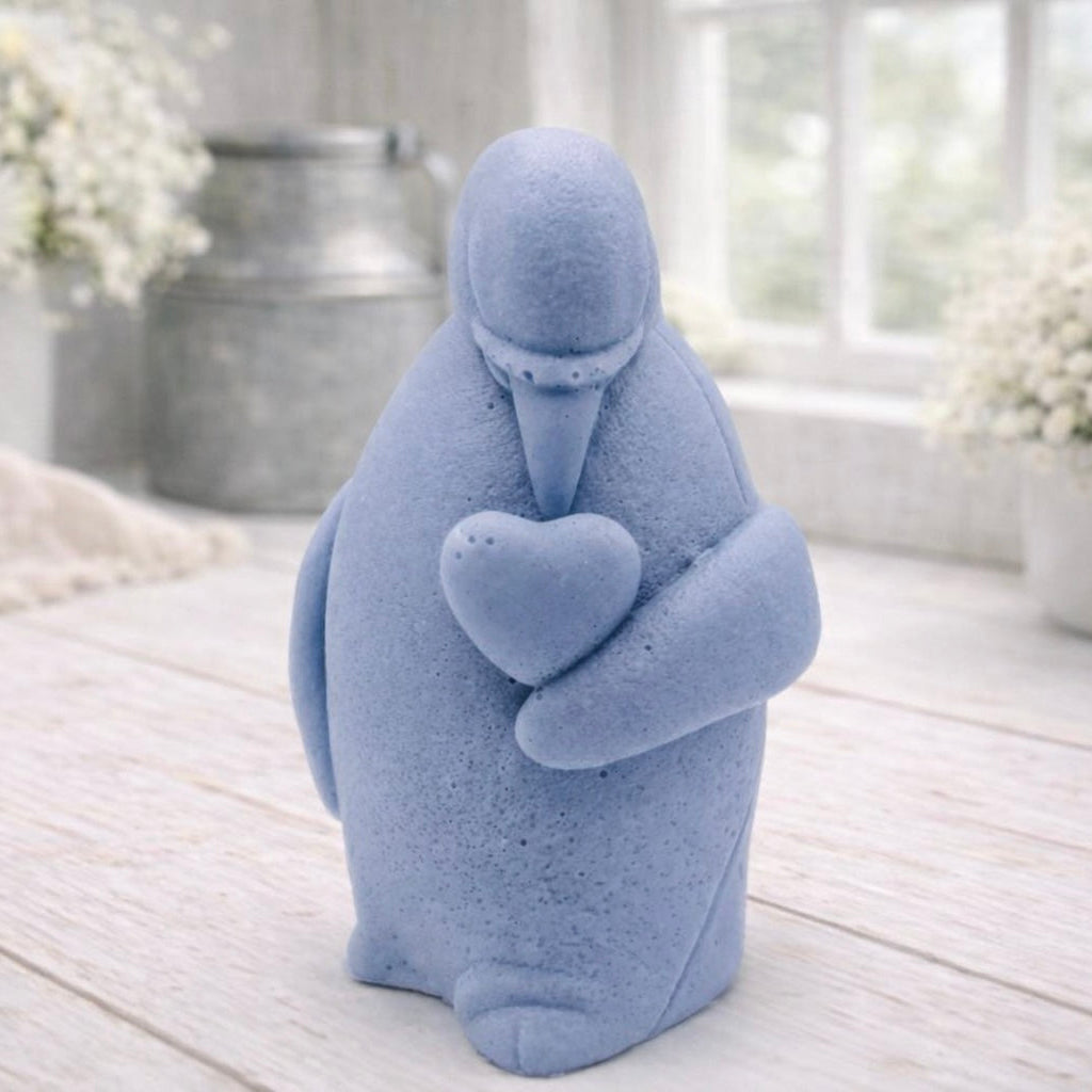 A 3D handmade vegan soap sculpted as a standing penguin tenderly cradling a heart against its chest, displayed on a light wooden surface in a bright farmhouse setting.