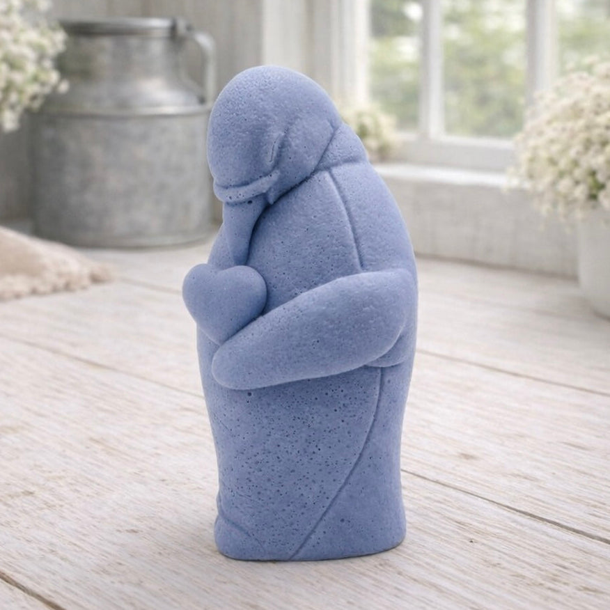 A 3D handmade vegan soap sculpted as a standing penguin tenderly cradling a heart against its chest, displayed on a light wooden surface in a bright farmhouse setting.