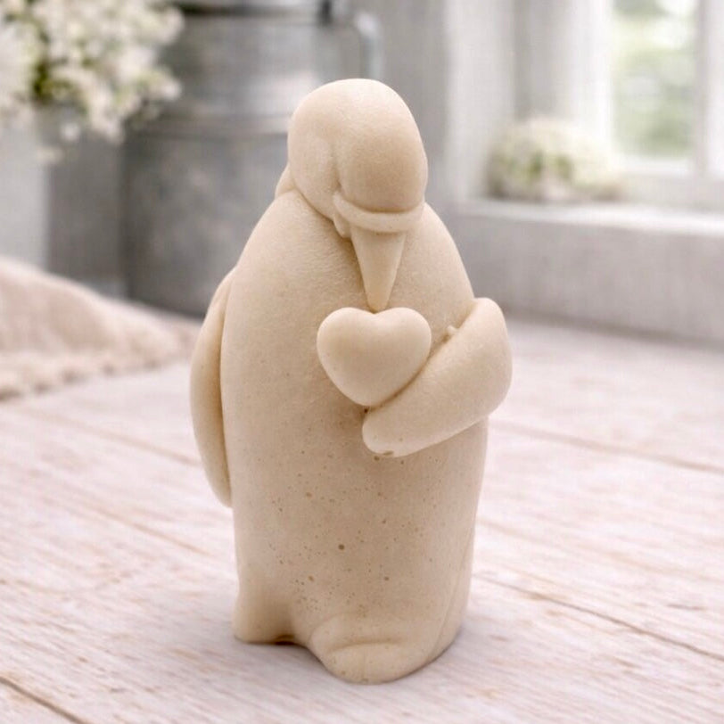A 3D handmade vegan soap sculpted as a standing penguin tenderly cradling a heart against its chest, displayed on a light wooden surface in a bright farmhouse setting.