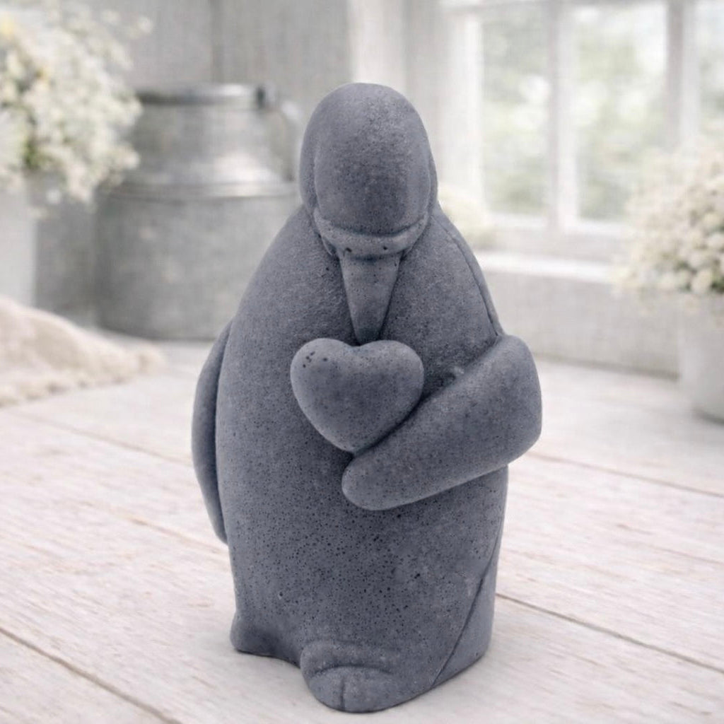 A 3D handmade vegan soap sculpted as a standing penguin tenderly cradling a heart against its chest, displayed on a light wooden surface in a bright farmhouse setting.