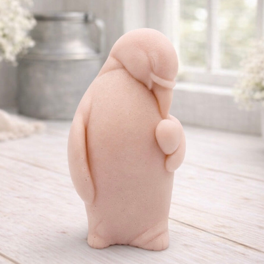 A 3D handmade vegan soap sculpted as a standing penguin tenderly cradling a heart against its chest, displayed on a light wooden surface in a bright farmhouse setting.
