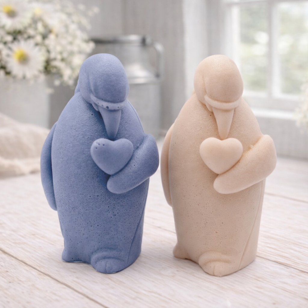 A 3D handmade vegan soap sculpted as a standing penguin tenderly cradling a heart against its chest, displayed on a light wooden surface in a bright farmhouse setting.