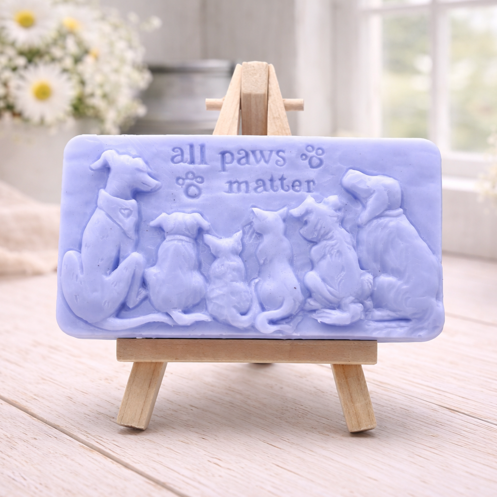 A rectangular handmade vegan soap bar gift, featuring a detailed relief carving of several dogs and a cat sitting together, with the words "all paws matter" and two paw prints embossed at the top, displayed on a wooden easel. Made in Blackpool. 