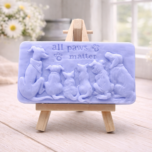 A rectangular handmade vegan soap bar gift, featuring a detailed relief carving of several dogs and a cat sitting together, with the words "all paws matter" and two paw prints embossed at the top, displayed on a wooden easel. Made in Blackpool. 