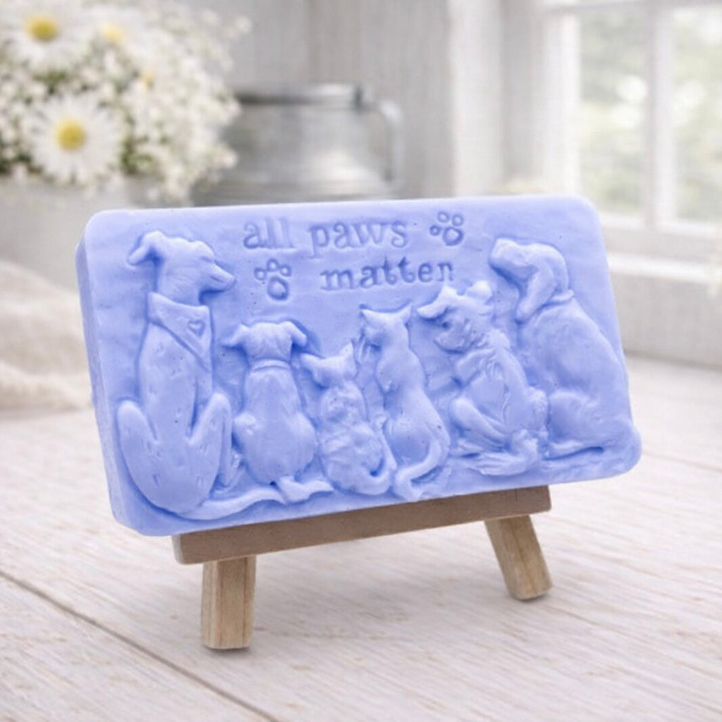 A rectangular handmade vegan soap bar gift, featuring a detailed relief carving of several dogs and a cat sitting together, with the words "all paws matter" and two paw prints embossed at the top, displayed on a wooden easel. Made in Blackpool. 