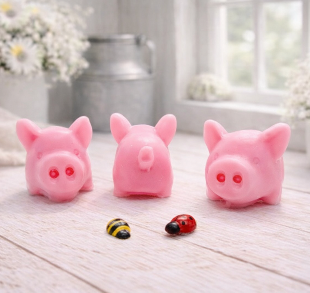 A set of three handmade vegan soaps sculpted into cute pigs with round snouts and floppy ears, displayed with a small decorative bee and ladybird on a white wooden surface.
