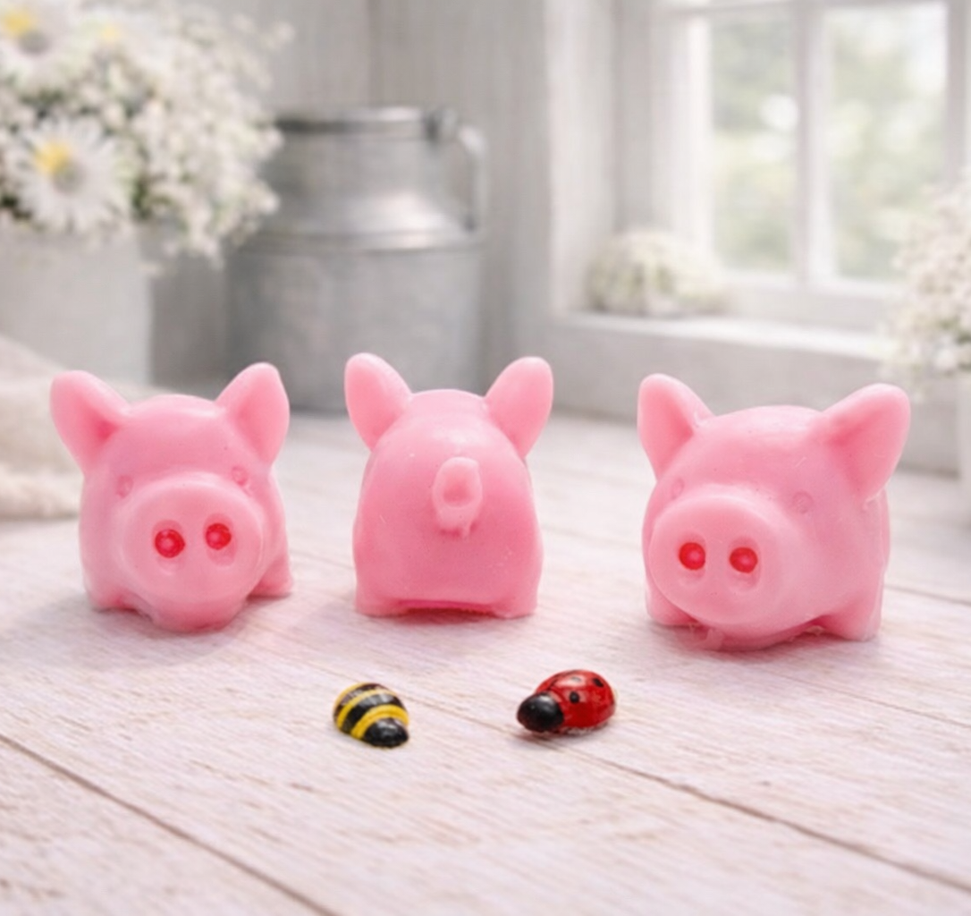 A set of three handmade vegan soaps sculpted into cute pigs with round snouts and floppy ears, displayed with a small decorative bee and ladybird on a white wooden surface.