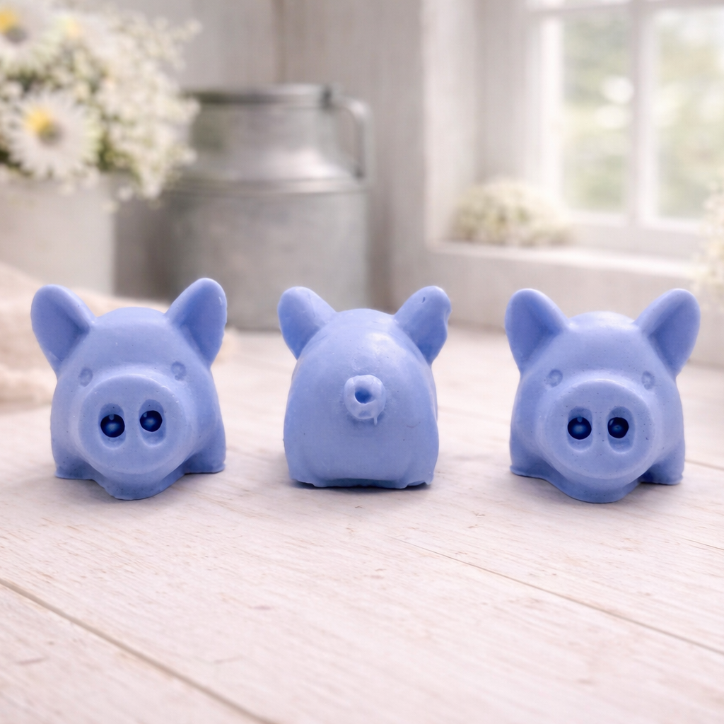 A set of three handmade vegan soaps sculpted into cute pigs with round snouts and floppy ears, displayed with a small decorative bee and ladybird on a white wooden surface.