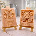 Two rectangular handmade vegan soaps displayed on miniature wooden easels; the left soap features a circular butterfly relief with the words "Embrace Your Dreams," and the right soap features a sunflower relief with the words "Life is Good".