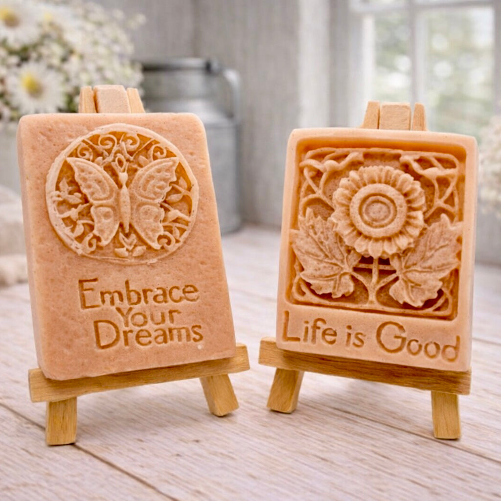 Two rectangular handmade vegan soaps displayed on miniature wooden easels; the left soap features a circular butterfly relief with the words "Embrace Your Dreams," and the right soap features a sunflower relief with the words "Life is Good".