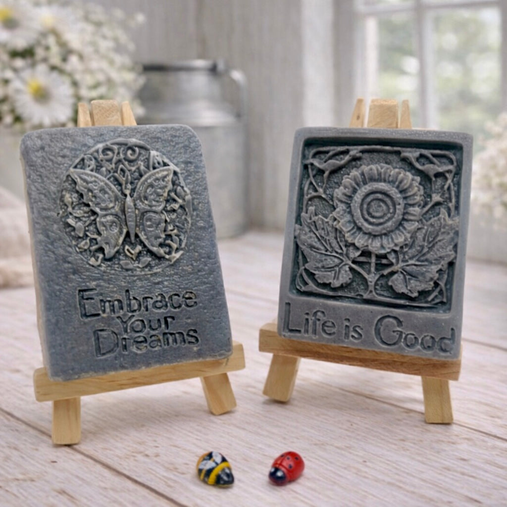 Two rectangular handmade vegan soaps displayed on miniature wooden easels; the left soap features a circular butterfly relief with the words "Embrace Your Dreams," and the right soap features a sunflower relief with the words "Life is Good".