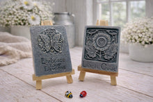 Load image into Gallery viewer, Two rectangular handmade vegan soaps displayed on miniature wooden easels; the left soap features a circular butterfly relief with the words "Embrace Your Dreams," and the right soap features a sunflower relief with the words "Life is Good".

