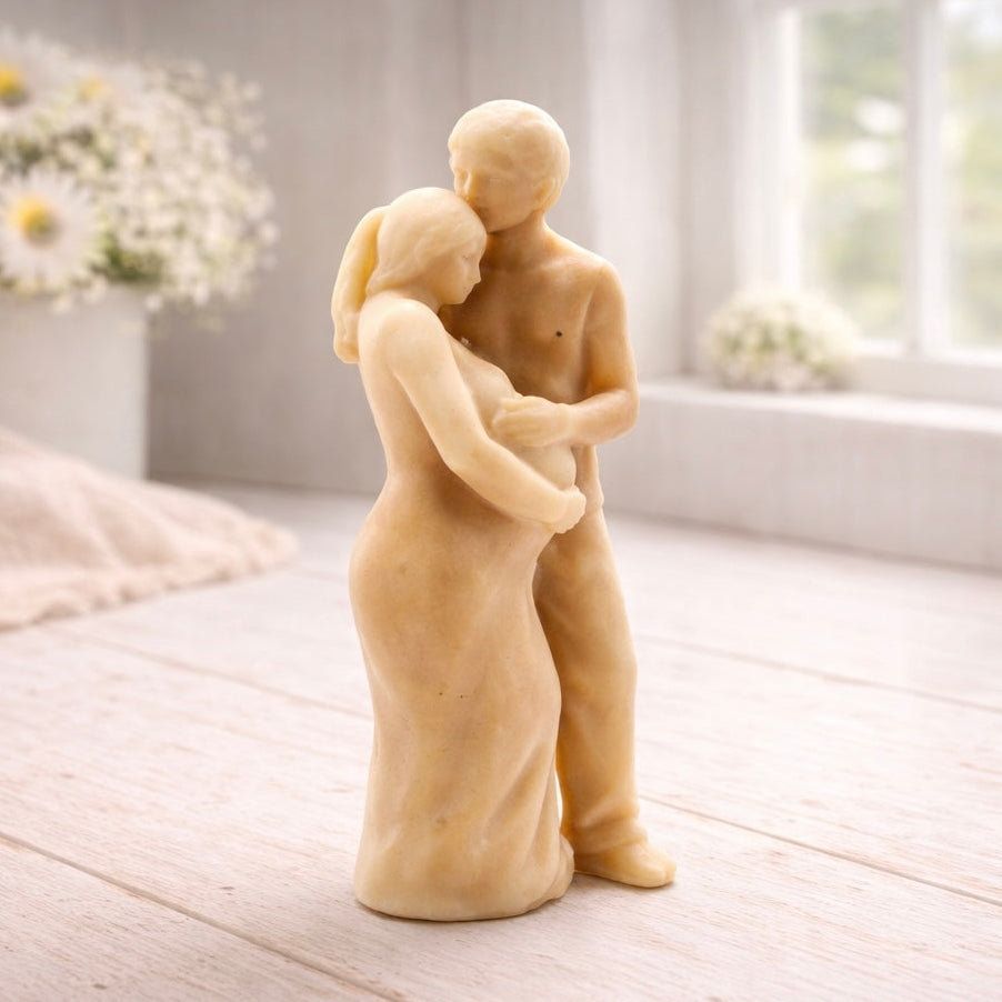 A 3D handmade vegan soap in a warm cream tone, sculpted into a standing figure of a pregnant couple; the partner stands behind the expectant mother, tenderly cradling her baby bump, displayed on a light wooden surface in a bright farmhouse setting.