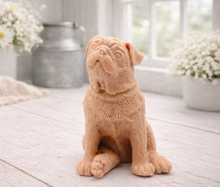 Load image into Gallery viewer, A detailed handmade vegan soap sculpted in the shape of a sitting pug dog with characteristic facial wrinkles and a tilted head, in a charcoal-grey tone, displayed on a light wooden surface.
