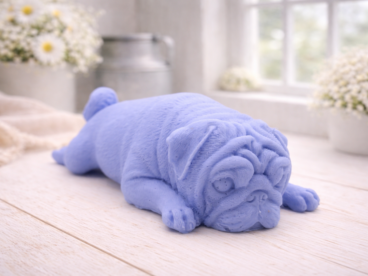 A detailed handmade vegan soap gift, sculpted in the shape of a pug dog lying flat on its stomach in a sleeping position, showing realistic skin folds and fur texture, displayed on a light wooden surface. Handmade in Blackpool 