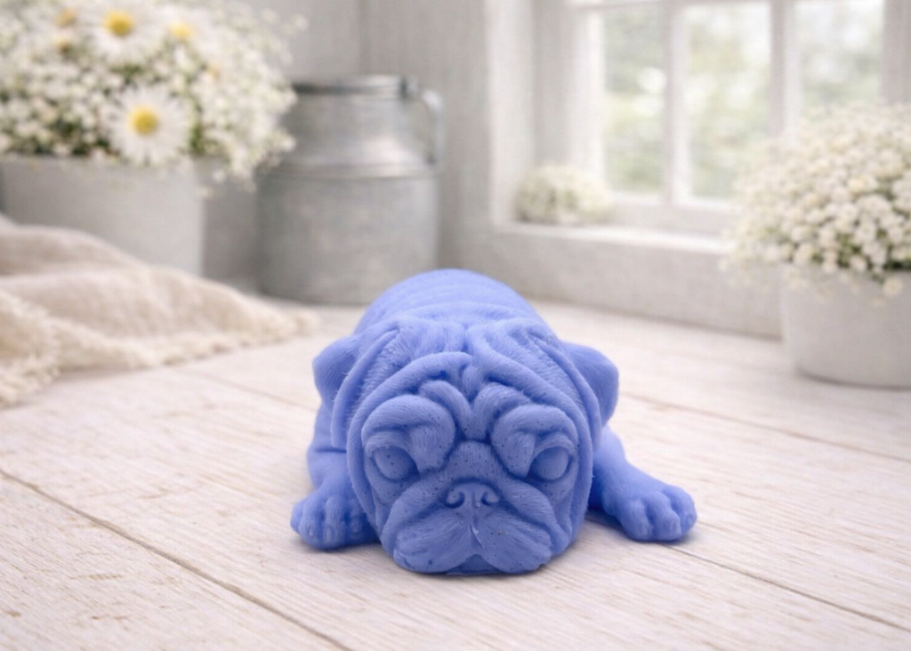 A detailed handmade vegan soap gift, sculpted in the shape of a pug dog lying flat on its stomach in a sleeping position, showing realistic skin folds and fur texture, displayed on a light wooden surface. Handmade in Blackpool 